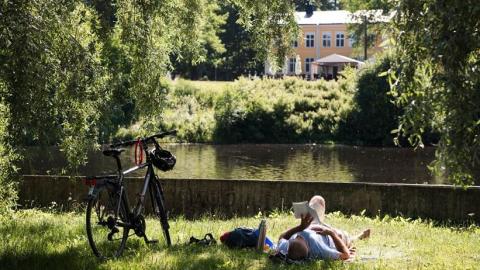 Man liger I gräset bredvid sin cykel, framför en å med ett stort hus i bakgrunden och läser