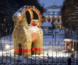 Gävlebocken i halm står i en inhägnad med en gul byggnad i bakgrunden en härlig vinterkväll.