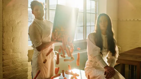 Två personer tittar in i kameran medan en av dem häller ut en låda med morötter