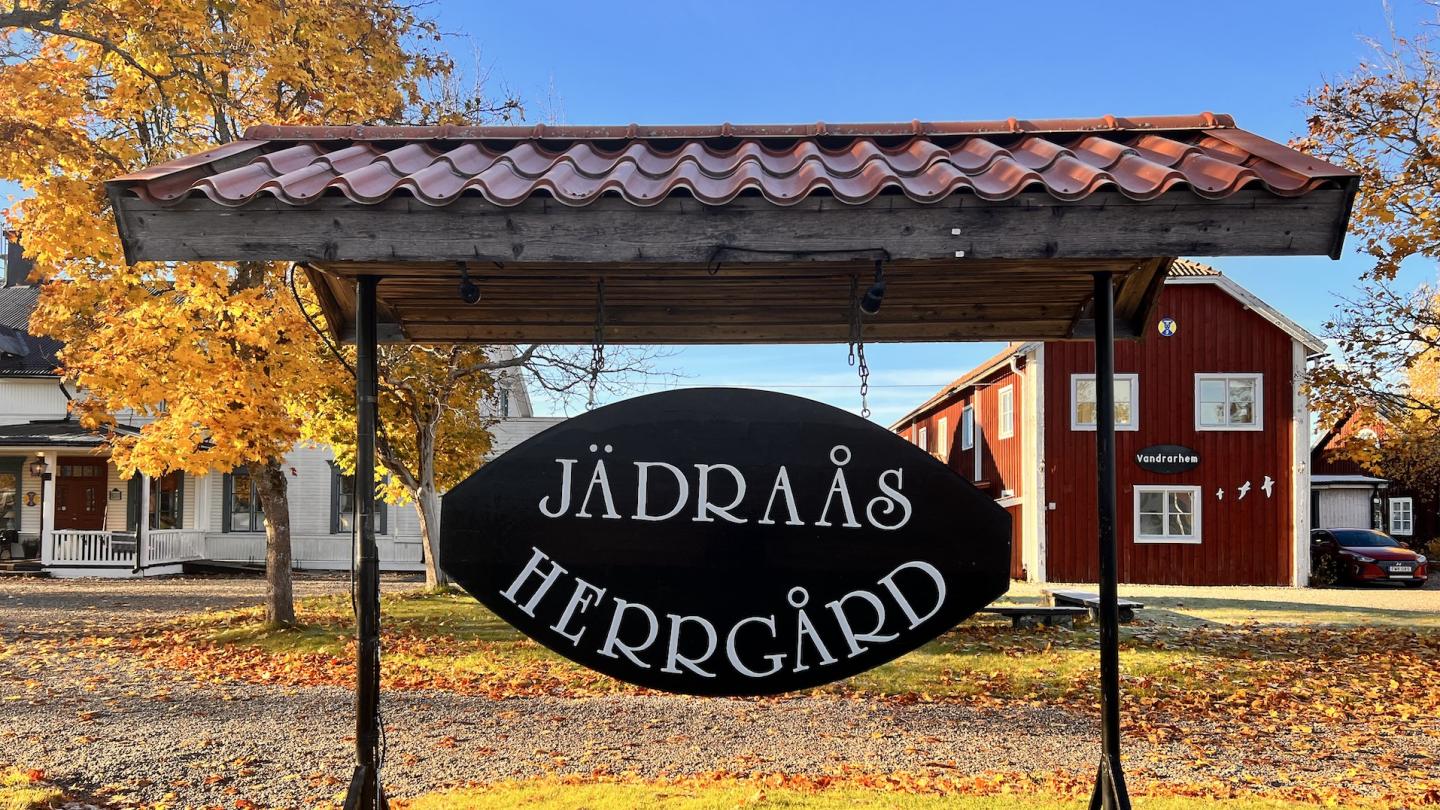 Stor skylt med texten Jädraås Herrgård med två byggnader i bakgrunden en fin dag i oktober